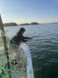 A young angler is actively reeling in a fish from the side of a boat on a serene lake, surrounded by lush greenery.