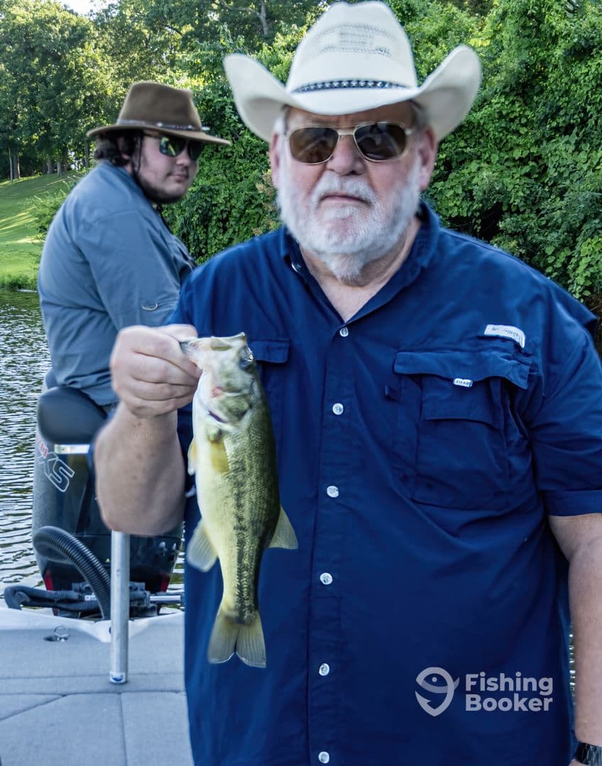 An angler proudly displaying a large Largemouth Bass while fishing on a serene lake, with another fisherman in the background.