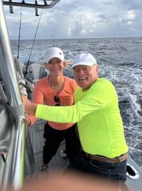A cheerful couple enjoying their time on a fishing boat, with a scenic ocean backdrop and fishing gear visible.