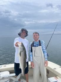 Two anglers proudly displaying a large Striped Bass while fishing on a boat, showcasing a successful day on the water.
