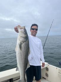 An angler proudly displaying a large Striped Bass while fishing on a boat, showcasing a successful day on the water.