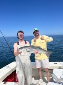 Two anglers proudly displaying a large Striped Bass while fishing on a boat in open water.