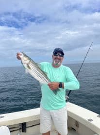 An angler proudly displaying a large Striped Bass while fishing on a boat, showcasing a successful day on the water.