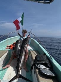An angler proudly displaying a large Marlin aboard a small boat in open waters, with the Mexican flag waving in the background.