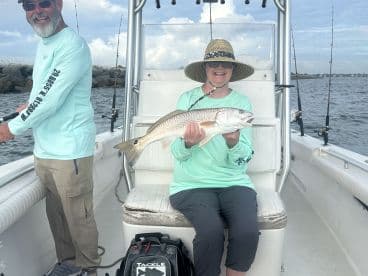 An adult angler proudly displaying a Redfish while seated on a boat, with another person actively fishing in the background.