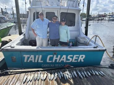 A group of anglers posing by the 'Tail Chaser' boat, showcasing their catch of Spanish Mackerel arranged on the dock after a successful fishing trip.
