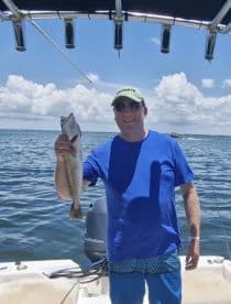 An angler proudly displaying a Speckled Trout while fishing on a sunny day, showcasing a vibrant blue sky and calm waters.