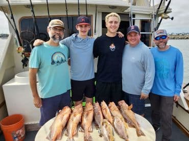 A group of four anglers proudly display their catch of several rockfish on the deck of a charter boat, showcasing a successful fishing trip.
