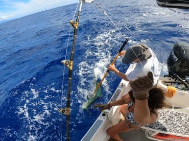 A young angler actively reeling in a fish while another person assists on a fishing boat in open waters.