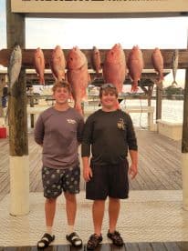 Two young anglers proudly posing in front of a catch board displaying several Red Snapper after a successful fishing trip.