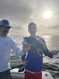 A young angler proudly displaying a Black Sea Bass while fishing on a boat, with a scenic ocean backdrop.