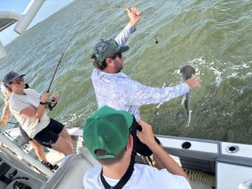 A group of anglers actively fishing on a boat, with one person reeling in a shark while others assist and capture the moment.