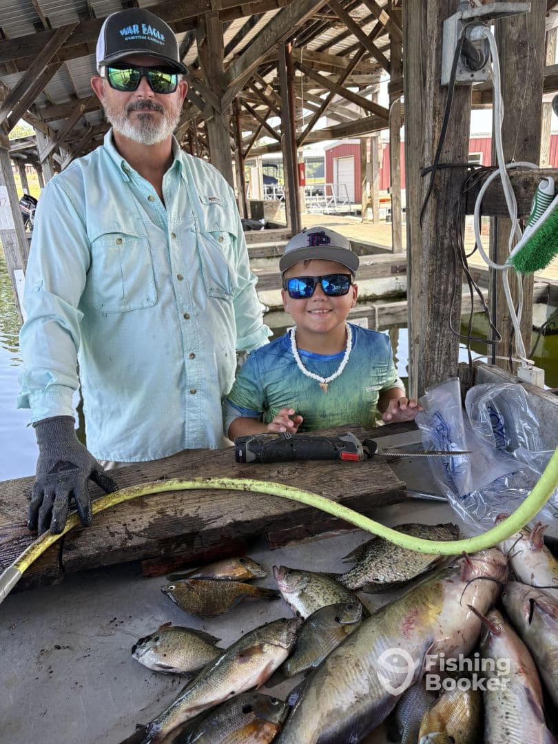 A proud young angler and an adult display a variety of freshly caught fish, including panfish and catfish, on a cleaning table at a dock.
