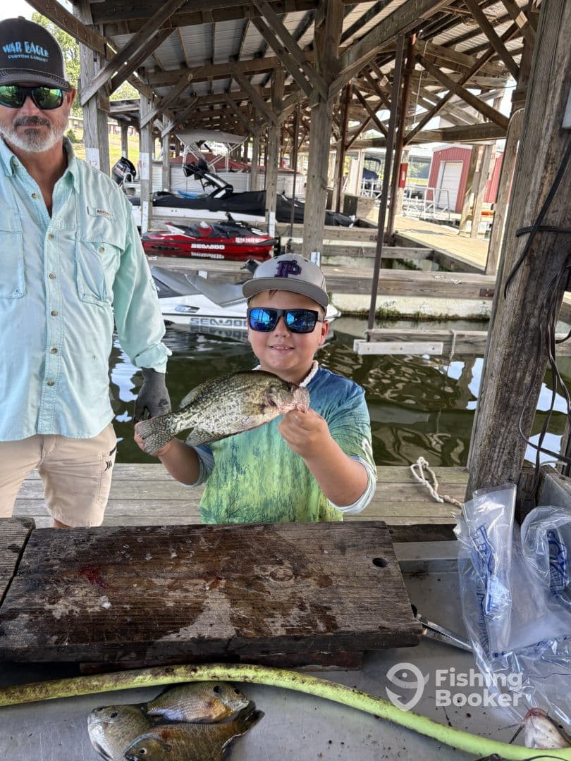 A young angler proudly displaying a large Crappie while standing at a dock, with boats visible in the background.
