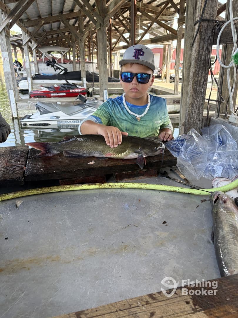 A young angler proudly displaying a catfish at a dock, surrounded by boats, showcasing a fun fishing experience.