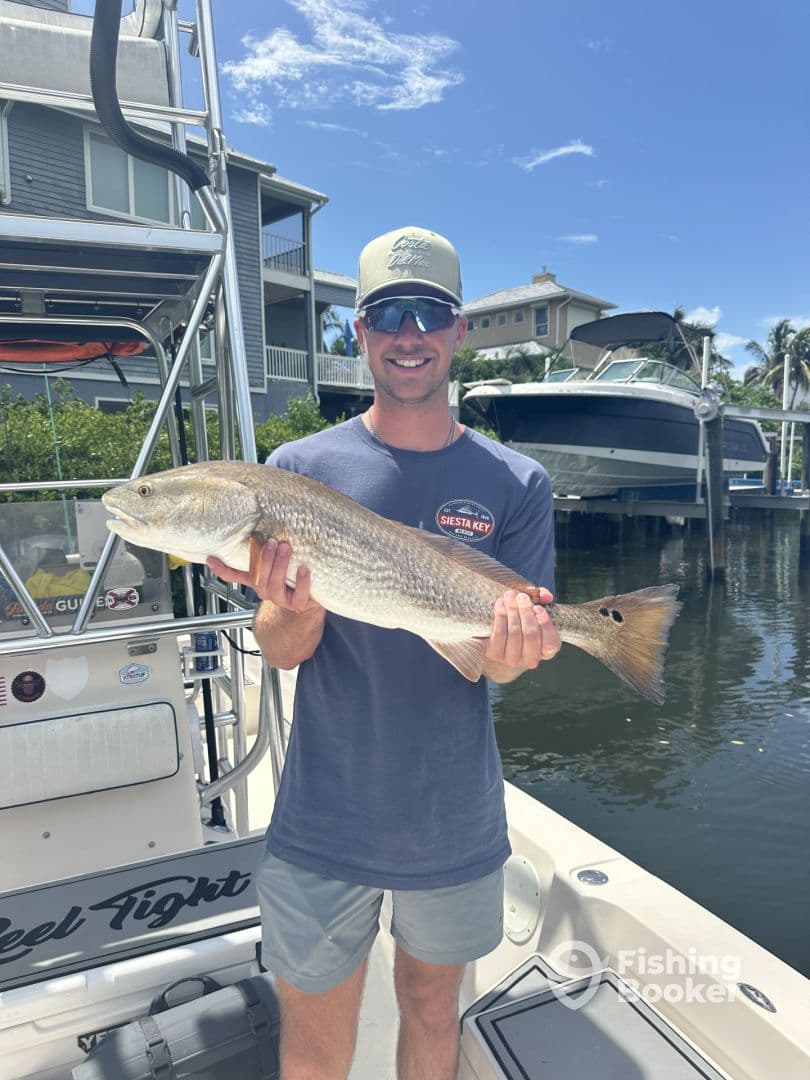Angler proudly displaying a large Redfish while standing on a boat docked in a residential area, showcasing a sunny day on the water.