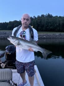 Angler holding a large Striped Bass while standing on a boat, showcasing a successful catch during an evening fishing trip.