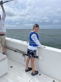 A young angler wearing a life jacket enjoys the thrill of fishing on a boat, with an adult preparing to cast a line in the background.