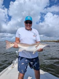 Angler proudly displaying a large Redfish while standing on the deck of a boat, with a scenic backdrop of clouds and water.