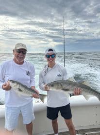 A proud young angler and an adult holding two striped bass while on a fishing trip, with a scenic ocean backdrop.