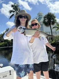 Two young anglers proudly displaying a large Snook on a sunny day while fishing from a boat in a scenic waterway.