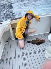 A young angler in a yellow rain jacket kneeling on the deck of a boat, proudly displaying a large Grouper caught during a fishing trip.