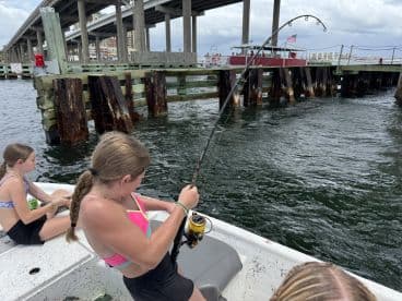 Two young anglers on a boat, with one actively reeling in a fish while the other observes, set against a backdrop of a dock and bridge.