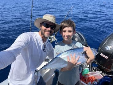 A young angler and an adult proudly displaying a large Bonito while fishing on a boat in deep blue waters.