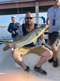 A proud angler displaying a large Snook while seated on a boat, with a young boy in the background enjoying the fishing experience.