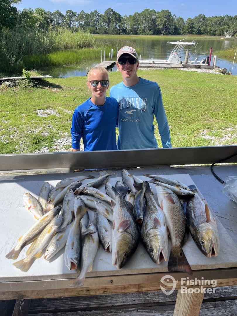 Two young anglers proudly posing with a large catch of various fish, including speckled trout and redfish, displayed on a cleaning table after a successful fishing trip.