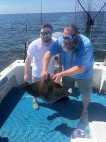 Two anglers proudly displaying a large stingray on a fishing boat, enjoying a sunny day on the water.
