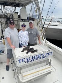 A family poses on the deck of the 'Teacher's Pet' charter boat, showcasing their catch of several flounder after a successful fishing trip.