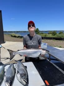 An angler proudly displaying a large Salmon at a scenic lakeside location, with several other fish laid out on a table.