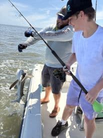 A young angler and an adult are reeling in a shark while fishing on a boat, showcasing an exciting moment on the water.