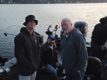 Three anglers relaxing inside a fishing boat at dusk, with fishing rods ready for action on a calm lake.