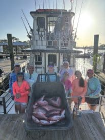 A group of anglers proudly posing with a large catch of Snapper displayed in a wheelbarrow on the deck of a charter boat, showcasing a successful fishing trip.