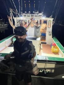 A young angler in a life jacket proudly displays his catch while two friends celebrate on the boat after a night fishing trip.