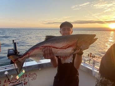 A young angler proudly displaying a large Lake Trout while fishing at sunset on a charter boat.
