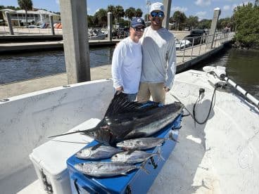 Two anglers proudly displaying a large Sailfish alongside several smaller fish on a boat after a successful fishing trip.