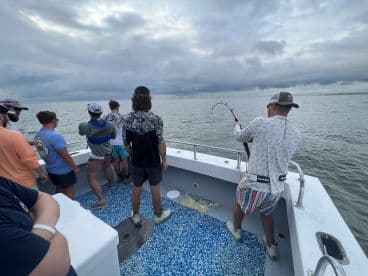 Group of anglers actively fishing on a charter boat, with one person reeling in a catch against a cloudy backdrop.