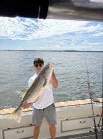 A young angler proudly displaying a large Striped Bass while fishing on a boat, with a scenic water backdrop.