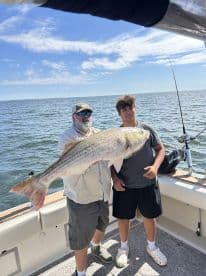 Two anglers proudly displaying a large Striped Bass aboard a fishing boat, enjoying a successful day on the water.