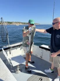 A young angler proudly displays a large Bluefish while fishing on a boat, with a scenic coastal backdrop.