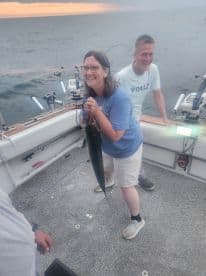 A happy angler holding a sizable fish aboard a fishing boat during an evening trip, with a scenic ocean backdrop.