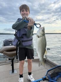 Young angler proudly holding a striped bass while fishing from a boat, showcasing a successful catch on a sunny day.