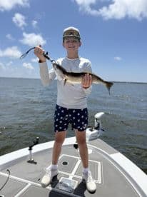 A young angler proudly displaying a Speckled Trout while standing on the deck of a boat under a bright blue sky.