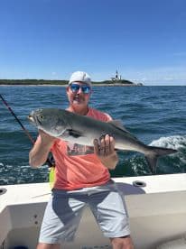 An angler proudly displays a large Bluefish while fishing off the coast, with a scenic lighthouse in the background.