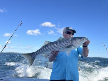 An angler proudly displaying a large Striped Bass while fishing on a boat, showcasing a beautiful day on the water.