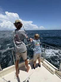 A young boy and an older companion are actively fishing on a boat, enjoying a sunny day on the water with a clear blue sky.