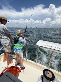 A young angler in a life jacket is reeling in a fish while fishing on a boat in a sunny coastal environment.